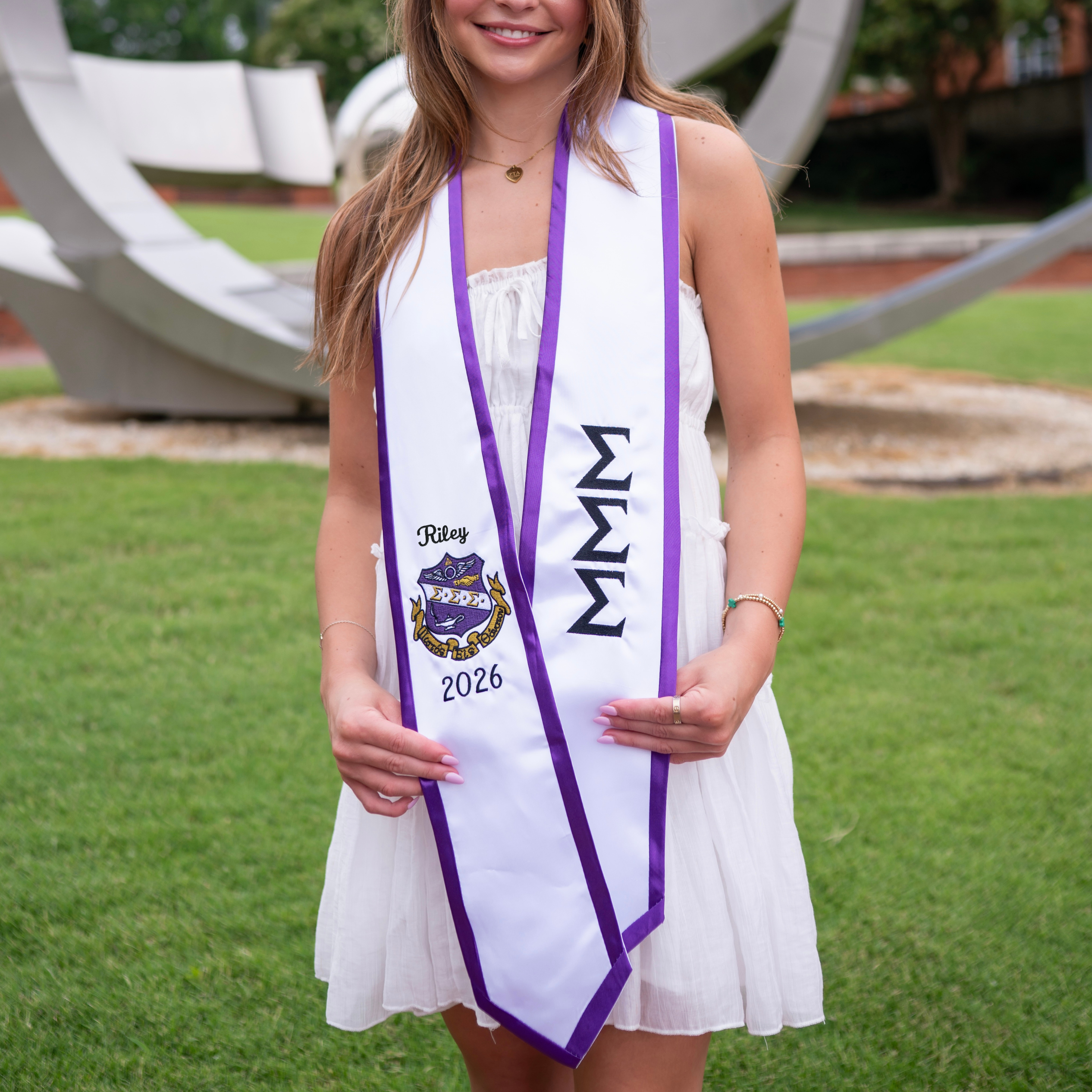 Graduate wearing a white gown with a purple and black stole Sigma Sigma Sigma or Tri Sigma stole outdoors on a grassy area.